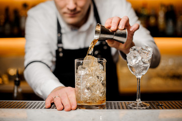 Barman dressed in a white shirt pouring alcoholic drink into a glass with ice cubes