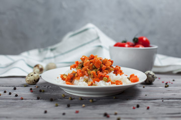 tasty rice with stewed carrots, peas and onions on a white plate, quail eggs, bowl with cherry tomatoes, pepper and towel on a light wooden table on a light gray background