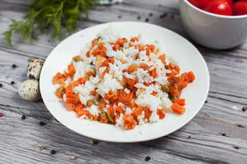 delicious rice with stewed carrots, peas and onions on a white plate, quail eggs, bowl with cherry tomatoes, towel, pepper and dill on a light wooden table