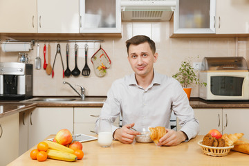 Handsome caucasian young business man in plaid shirt having breakfast, sitting at table, eating cereals and croissant with milk on light kitchen. Healthy lifestyle. Cooking at home. Prepare food.