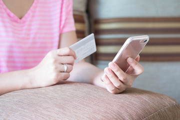 Close up woman hands holding credit card and using cell phone for online shopping on sofa in the living room of the house.