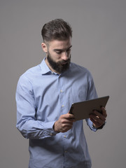 Standing young adult bearded business man holding and watching at tablet computer over gray studio background. 