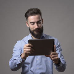 Young happy bearded man holding and reading e-book from touch pad tablet computer over gray studio background. 
