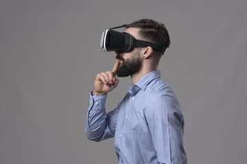 Minimalism profile view of young business man wearing vr glasses with finger over lips looking away at copyspace over gray studio background. 