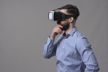 Young adult smart casual businessman wearing head mounted vr headset looking away at copyspace and thinking over gray background. 