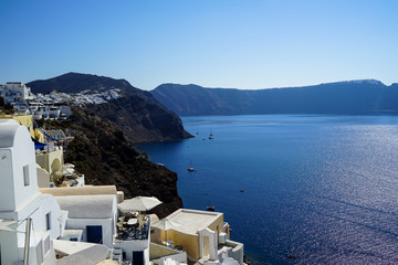 Panoramic view of blue Aegean sea, sailing ships and ocean water reflection from Oia village with white buildings cityscape along island mountain and blue sky background