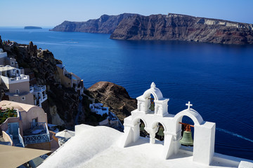 Panoramic view of beautiful blue Aegean sea and caldera from Oia village with white church foreground, buildings along island mountain and blue sky background