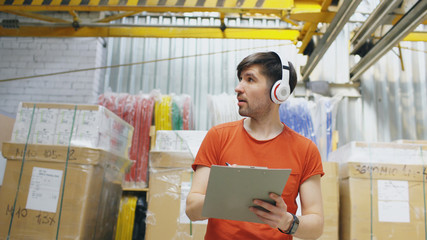 Happy young worker in industrial warehouse listening to music during work. Man in headphones have fun at workplace.