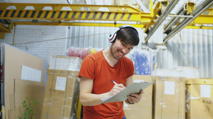 Happy young worker in industrial warehouse listening to music and dancing during work. Man in headphones have fun at workplace.