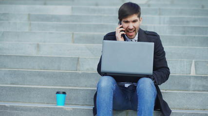 Young business man with laptop computer having stress after phone call and sitting on stairs in street. Businessman having deal problems at work concept