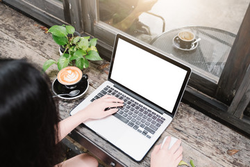 Mockup image of business woman using and typing on laptop with blank white screen and coffee cup on glass table in modern loft cafe, Soft focus on vintage wooden table.