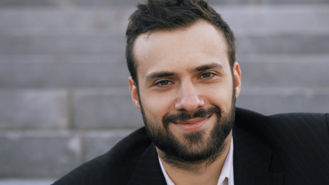 Close up portrait of bearded young businessman smiling and looking into camera in street outdoors