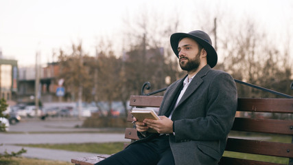 Fototapeta premium Creative young writer man in hat and coat write notes for his future book with pen in notebook sitting on city street bench at park