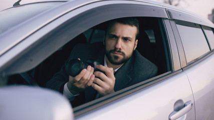 Young private detective man sitting inside car and photographing with dslr camera