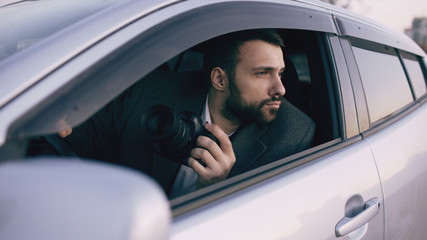 Young private detective man sitting inside car and photographing with dslr camera