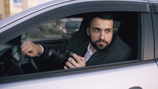 Young private detective man sitting inside car and photographing with dslr camera