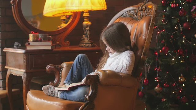 beautiful little girl is reading a book while sitting in a chair near a festive Christmas tree
