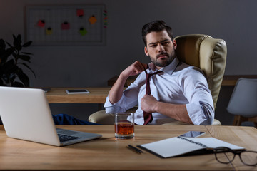 tired businessman loosen tie and sitting at table with glass of whiskey