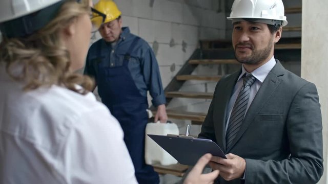 Slowmo of Asian businessman with clipboard standing in unfinished building and discussing work with female construction project manager in hard hat, then talking to male laborer in blue overalls