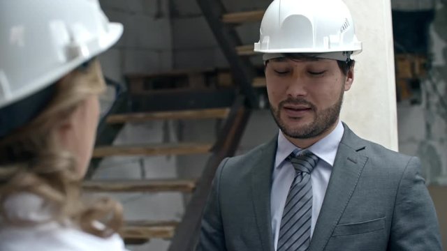Over the shoulder shot of Asian businessman in hard discussing work with female construction project manager while inspecting unfinished building; male laborer in blue overalls working in background