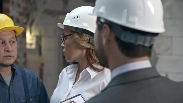 PAN of serious female construction coordinator in glasses and hard hat discussing work with unrecognizable businessman and cheerful mature foreman