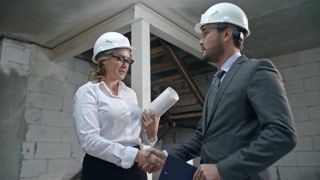 Medium shot of female construction contractor in hard hat holding blueprints and shaking hands with Asian businessmen signing documents on clipboard, then smiling and discussing work