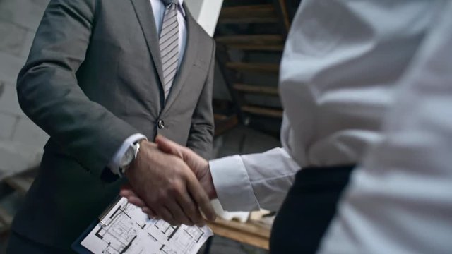 Tilt down of Asian construction engineer wearing suit and hard hat holding clipboard with layout plan and shaking hands with unrecognizable businesswoman