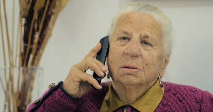 Close-up of an old granny of eighty-three years old talking to her friend on a phone while sitting on a chair in the room.