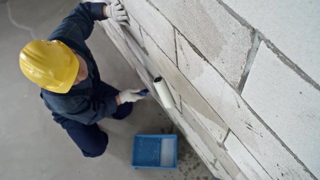 Top view of mature Asian construction painter in hard hat holding paint roller putting priming coat on brick wall in unfinished building