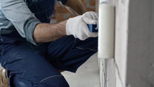 Tilt up of concentrated male Asian worker in blue overalls putting priming coat on brick wall with paint roller