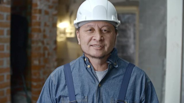 Tilt up of cheerful mature Asian builder in hard hat and overalls standing in unfinished building and smiling for camera