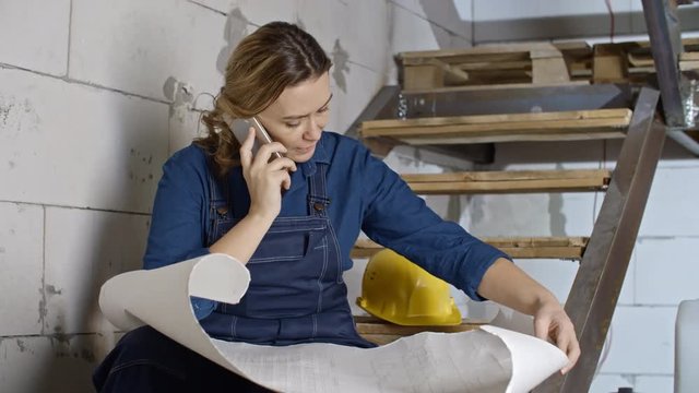 Tilt up of concentrated female engineer in blue overalls sitting on staircase in unfinished building and talking on mobile phone while inspecting blueprints