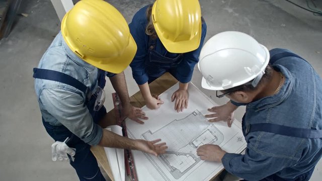 Directly above shot of male and female construction workers in hard hats standing and discussing floor plan draft