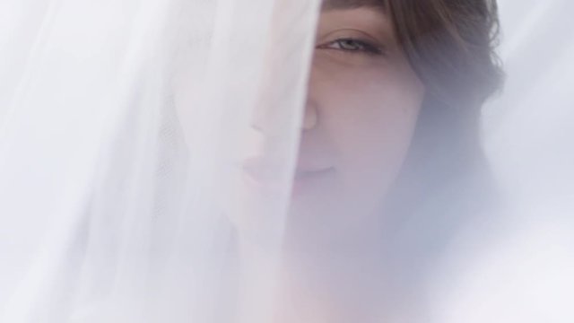 Slowmo of beautiful young woman standing behind white translucent tulle curtain and looking sensually into camera