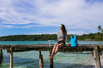 Brunette, young women, girl in striped jacket sits on the pier at the beach and look at sun. Better life. Thailand Asia. Sun glasses. Traveler. Waterproof bag