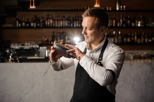 Barman in a white shirt and apron holding a shaker against the bar counter