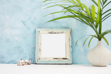 Square blue Photo frame mock up with green tropical plants in vaseand small wooden houses on shelf. Scandinavian style. Text space