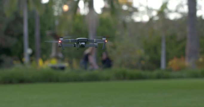 Remote controlled drone flying in a park setting in late afternoon. Visitors to park and other details out of focus in background. Slow motion, hand-held 4K.