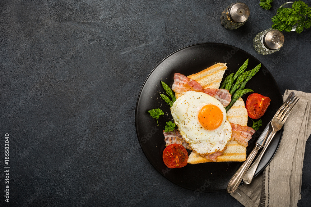 Wall mural Breakfast or lunch with Fried egg, bread toast, green asparagus, tomatoes and bacon on black plate. Top view. Copy space.