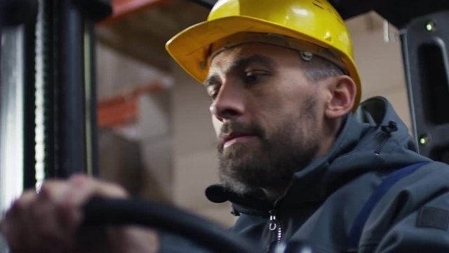 Low angle shot of bearded male worker in hard hat operating forklift in warehouse