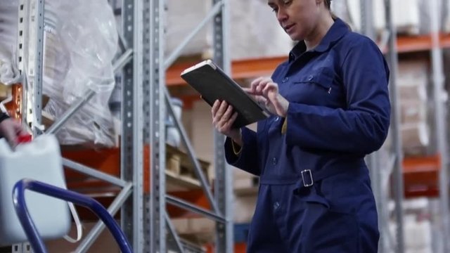 Tilt up of bearded warehouse worker in hard hat and uniform putting jerrycan in cart and discussing something with female colleague showing him something on tablet and pointing at rack shelves