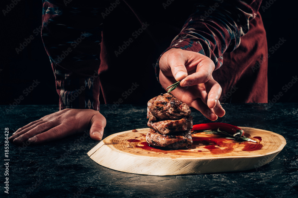 Wall mural close-up partial view of chef putting rosemary on grilled steaks with chili pepper and sauce on wooden board