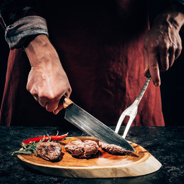 Close-up Partial View Chef In Apron With Meat Fork And Knife Slicing Gourmet Grilled Steaks With Rosemary And Chili Pepper On Wooden Board