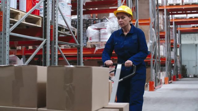 Tracking of female worker wearing uniform and hard hat pulling platform cart with cardboard boxes on it and walking through warehouse with rack shelves