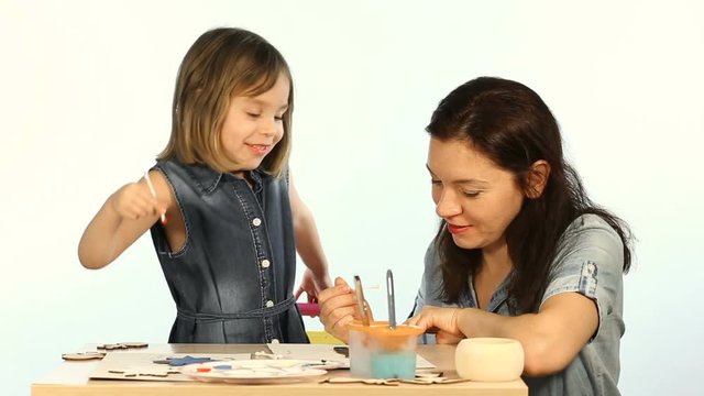 Shooting in the studio on a white background. A child, a funny girl, learns to draw. Mom shows you how to paint wooden New Year and Christmas figures. They are wearing a blue denim dress.