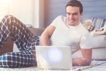 Pleasant time. Handsome positive relaxed man using his gadget for reading the emails while still wearing his pajamas