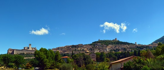 Fototapeta na wymiar Panoramic view of Assisi, from the plain to the hill of Assisi. On the left the imposing structure surmounted by the basilica. Clear blue sky with clouds. Photo of 13 August 2016 11:30