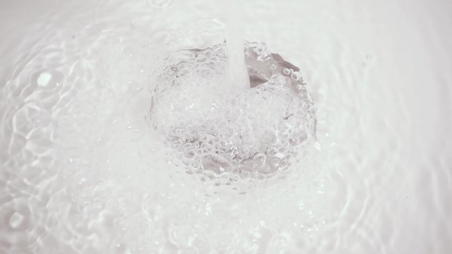 Slow motion bubbles of clear clear water in a silvery drain hole of a white bath or sink