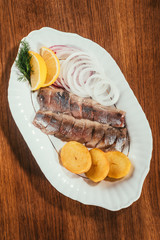 Top view of herring fish pieces with lemon, onion and potato on white plate on wooden table