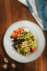 Top view of fresh salad with vegetables, eggs and salmon served on white plate with napkin on wooden table